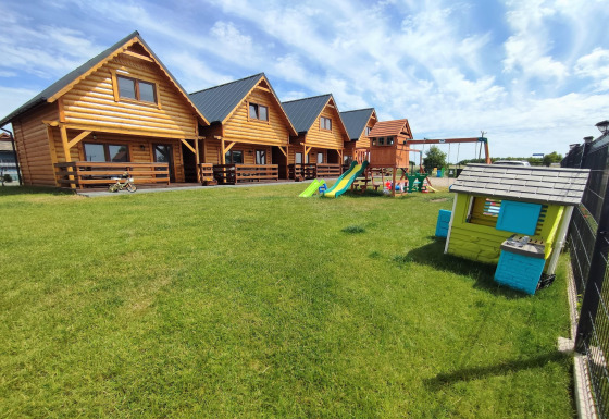 Cabañas de madera en Lazurowy Zakątek con zona infantil y césped verde bajo un cielo azul claro.