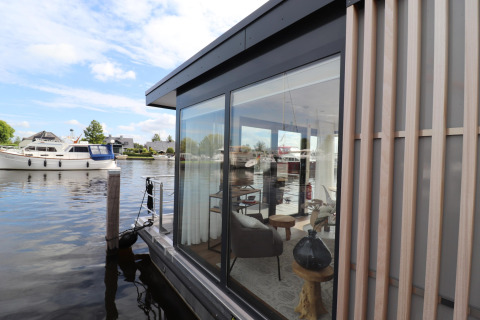 Zicht op woonboot «Refugio» in Nederland met glazen muren, modern interieur en boten op het water.
