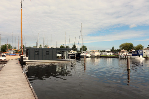 Houseboat Refugio moderne amarré dans un port avec voiliers et bateaux à moteur sous un ciel nuageux