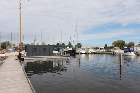 Moderne woonboot Refugio aangemeerd in een jachthaven met zeilboten en motorboten op een bewolkte dag