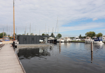 Houseboat Refugio moderne amarré dans un port avec voiliers et bateaux à moteur sous un ciel nuageux