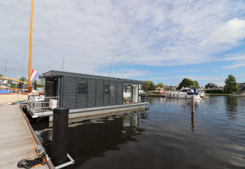 Péniche moderne Refugio amarrée à un quai aux Pays-Bas, entourée d'eau et de bateaux sous ciel bleu.