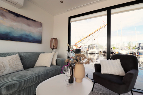 Stue på husbåd 'Refugio' i Holland med moderne sofa, stol, stort vindue og udsigt til bådehavn.