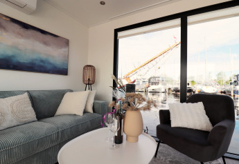 Stue på husbåd 'Refugio' i Holland med moderne sofa, stol, stort vindue og udsigt til bådehavn.