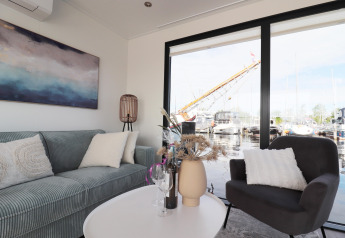 Soggiorno sulla houseboat 'Refugio' nei Paesi Bassi con arredo moderno e ampia finestra sul porto turistico.