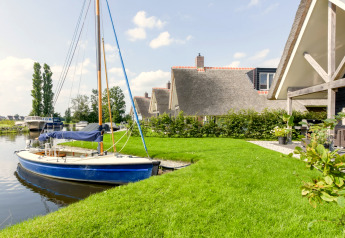 Chalet con tetto di paglia sull'acqua, barca a vela ormeggiata e prato verde curato in primo piano.