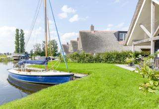 Chalet med stråtag ved vandet, sejlbåd fortøjet ved kajen og velplejet grøn græsplæne i forgrunden.