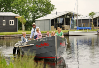 Een gezin vaart met een boot, 'euclides', langs moderne chalets in een groene, waterrijke omgeving.
