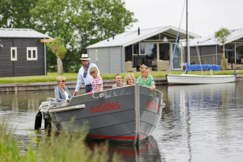 Eine Familie fährt mit einem Boot namens 'euclides' vor modernen Chalets und ruhigem Wasser entlang.