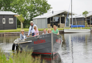 En familie sejler i en båd med navnet 'euclides' foran moderne chalet-hytter i grønne omgivelser.