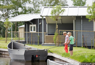 Deux enfants pêchent à côté d’un chalet au bord de l’eau, avec un bateau amarré dans un cadre verdoyant.