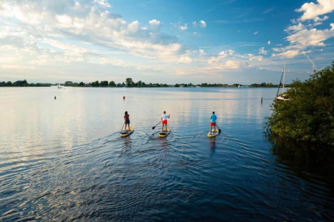 Tre persone fanno paddleboard su un lago tranquillo vicino a uno chalet sotto un cielo parzialmente nuvoloso.