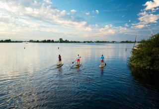 Three people paddleboarding on a calm lake near a chalet under a partly cloudy sky and bright sun.