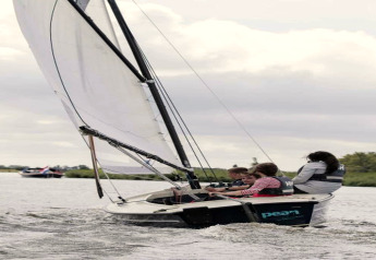 A group of people sails a small sailboat on a lake near a chalet, all wearing life jackets on a cloudy day.