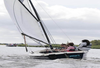 Vier Personen segeln gemeinsam in einem kleinen Segelboot auf einem See bei einem Chalet.