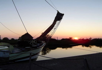 Foto di una barca vicino a uno chalet, ormeggiata su un lago calmo al tramonto con riflessi sull’acqua.
