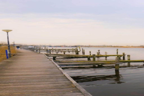 Wooden boardwalk beside calm water and empty docks, chalets visible in the distance under a cloudy sky.
