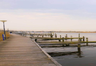 Wooden boardwalk beside calm water and empty docks, chalets visible in the distance under a cloudy sky.