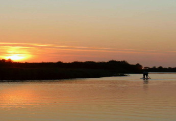 Beautiful sunset view from a chalet, with a person paddleboarding on the calm lake among peaceful scenery.