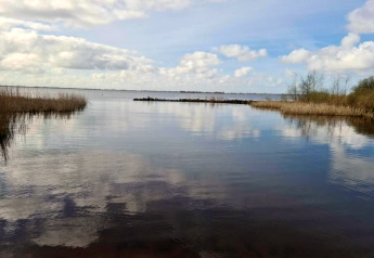 Zicht vanaf een chalet op een meer met blauwe lucht, weerspiegelende wolken en riet langs de kant.