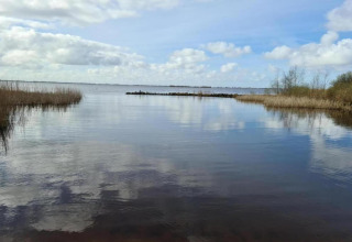 Blick auf einen ruhigen See bei einem Chalet, mit Wolkenspiegelung und Schilf am Ufer unter blauem Himmel.