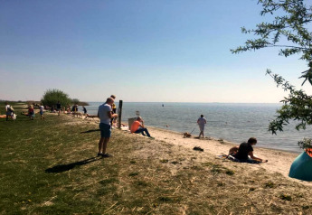 Persone che si rilassano davanti a un lago vicino a uno chalet, tra prato verde e spiaggia sabbiosa.