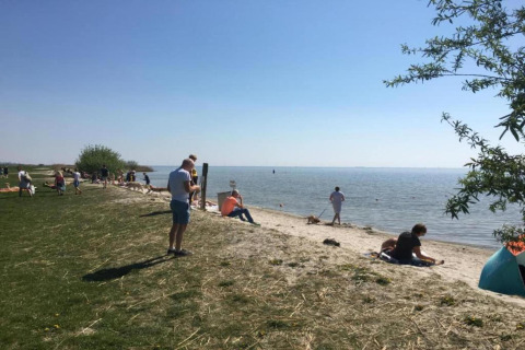 Personas disfrutando de un día soleado cerca de un chalet, sentadas en la playa de pasto y arena junto al lago.