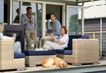 Vier volwassenen en een hond genieten van wijn op het terras van een chalet in het daglicht.