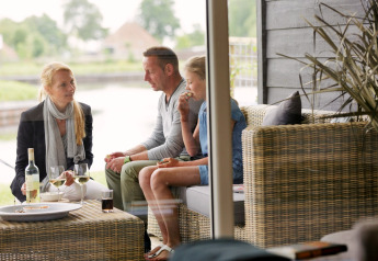 Tre persone chiacchierano e gustano vino su una terrazza di un chalet con vista sull’acqua.