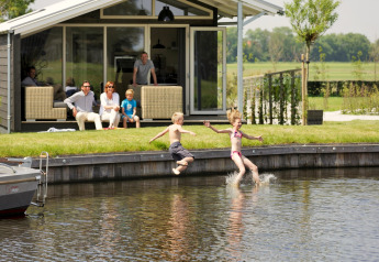 Two children jump into a lake near a chalet while their family watches and relaxes on the patio in summer.