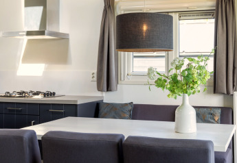 Moderne chaletkøkken og spiseområde med grå sofa, hvide borde, grønne planter og store vinduer.