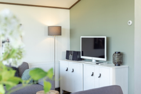 Living room in chalet Water park “Terkaple 1” in the Netherlands with TV, gray sofa, and white stand.