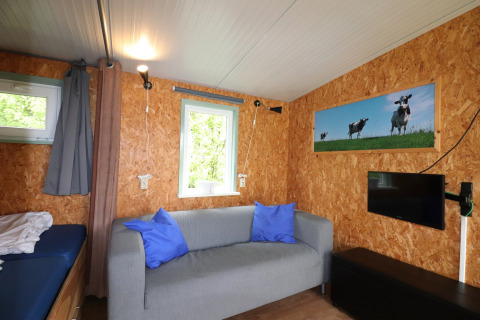 Salon lumineux du Cosy Cabin 1 aux Pays-Bas avec canapé gris, télé, fenêtre et photo de vaches.