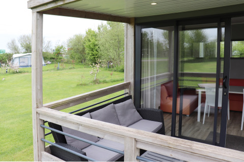 Terrazza esterna con divano e vista sul soggiorno della Terrace lodge 2 in un campeggio verde nei Paesi Bassi.