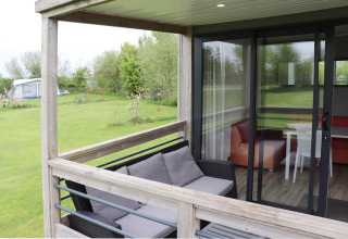 Terrazza esterna con divano e vista sul soggiorno della Terrace lodge 2 in un campeggio verde nei Paesi Bassi.