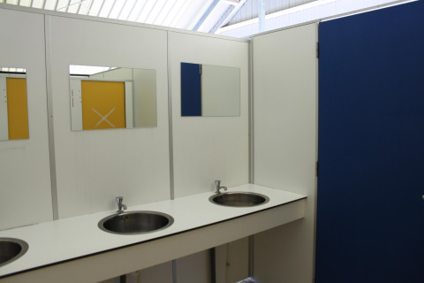Innenansicht eines öffentlichen Badezimmers mit zwei Waschbecken, Spiegeln und blauen sowie gelben Türen.