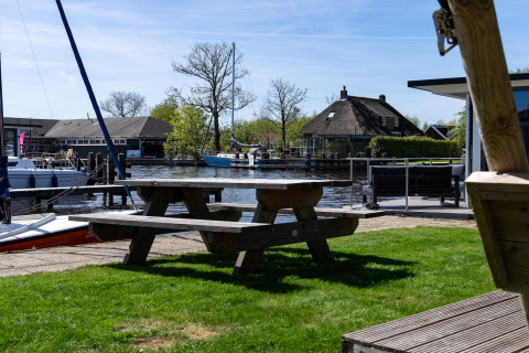 Blick von Safari Zelt Heeg in den Niederlanden mit Holztisch, Booten und traditionellem Haus am Wasser.