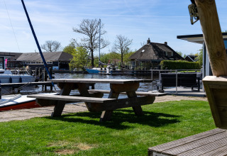 Blick von Safari Zelt Heeg in den Niederlanden mit Holztisch, Booten und traditionellem Haus am Wasser.