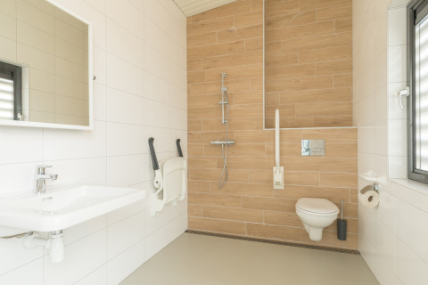 Rolstoelvriendelijke badkamer in een strandhuis met hangtoilet, inloopdouche, steunbeugels en groot raam.