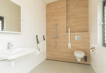Baño accesible en casa de playa con inodoro suspendido, ducha, barras de apoyo y una ventana grande.