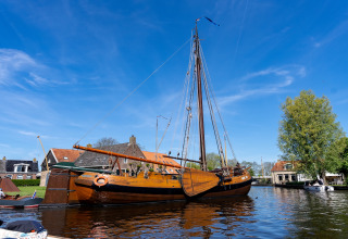 Traditionelles Segelboot liegt am Ufer nahe Safari Tent Heeg, mit Häusern im Hintergrund und blauem Himmel.