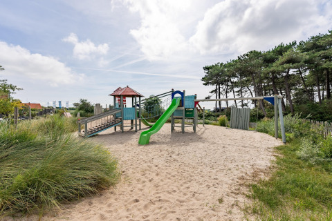 Speelplein met glijbaan en schommel op zand aan het strand, omringd door bomen en groen.