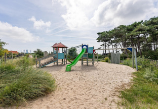 Speelplein met glijbaan en schommel op zand aan het strand, omringd door bomen en groen.