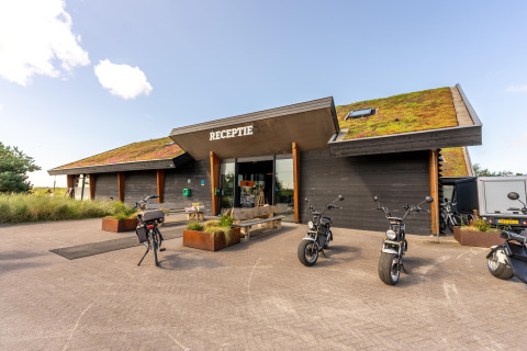 Afbeelding van de receptie van een strandhuis met groendak, fietsen en e-scooters buiten.