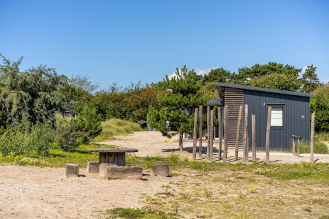 Knus strandhuisje met zand, bomen en een robuuste picknicktafel onder een blauwe hemel.
