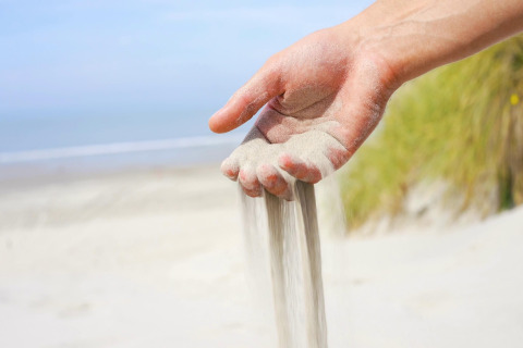 Een hand laat zand uitstrooien op het strand met zee en duingras, vlakbij een strandhuisje.