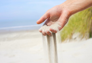 Een hand laat zand uitstrooien op het strand met zee en duingras, vlakbij een strandhuisje.