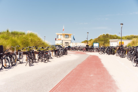 Veel fietsen staan geparkeerd bij een strandhuisje en redderspost, omringd door zandduinen en gras.