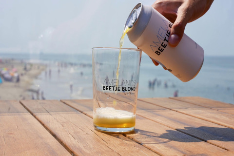 Een hand giet blond bier in een glas op een houten tafel met strand en zee zichtbaar op de achtergrond.