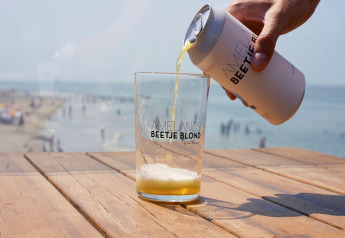 Una mano vierte cerveza rubia en un vaso sobre una mesa de madera con vista a la playa y al mar al fondo.
